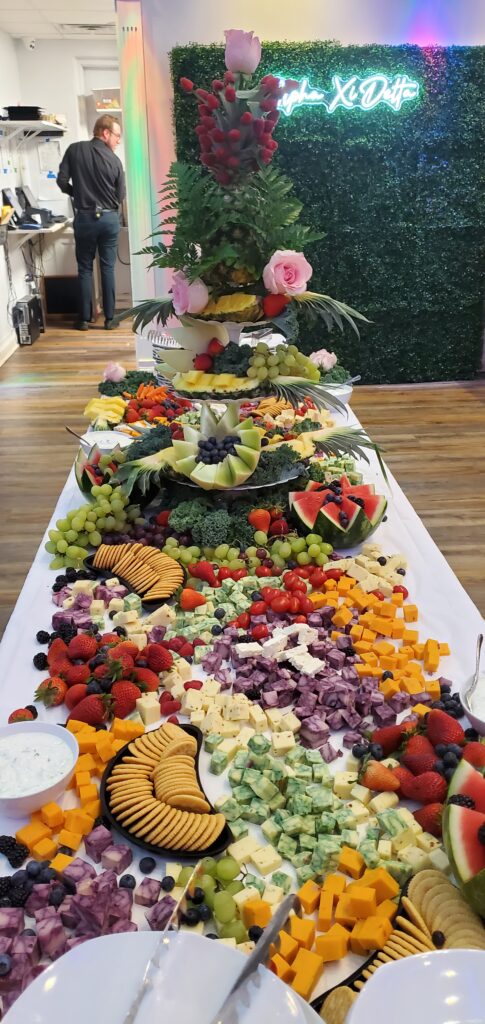 A vibrant charcuterie table, perfect for receptions or a ceremony in Bowling Green KY, displays assorted cheeses, crackers, fruits, and vegetables with a fruit and flower centerpiece. A neon sign and lush greenery wall complete the scene.
