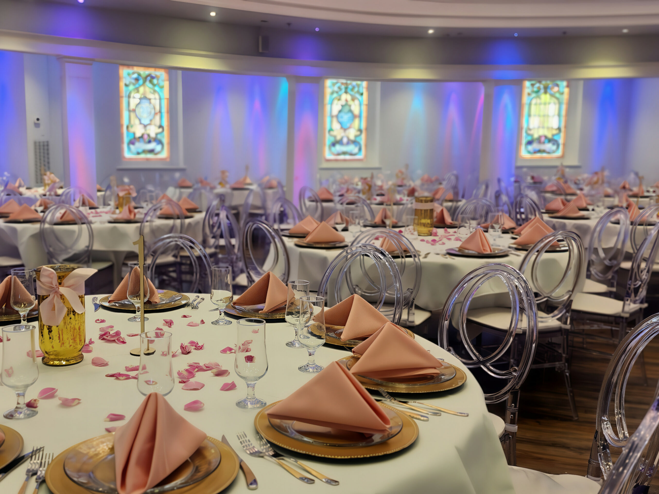 A banquet hall in Bowling Green KY set for a special event, featuring round tables with white tablecloths, pink folded napkins, gold plates, clear chairs, scattered rose petals—an ideal wedding venue for rehearsal dinners. Stained glass windows complete the look.