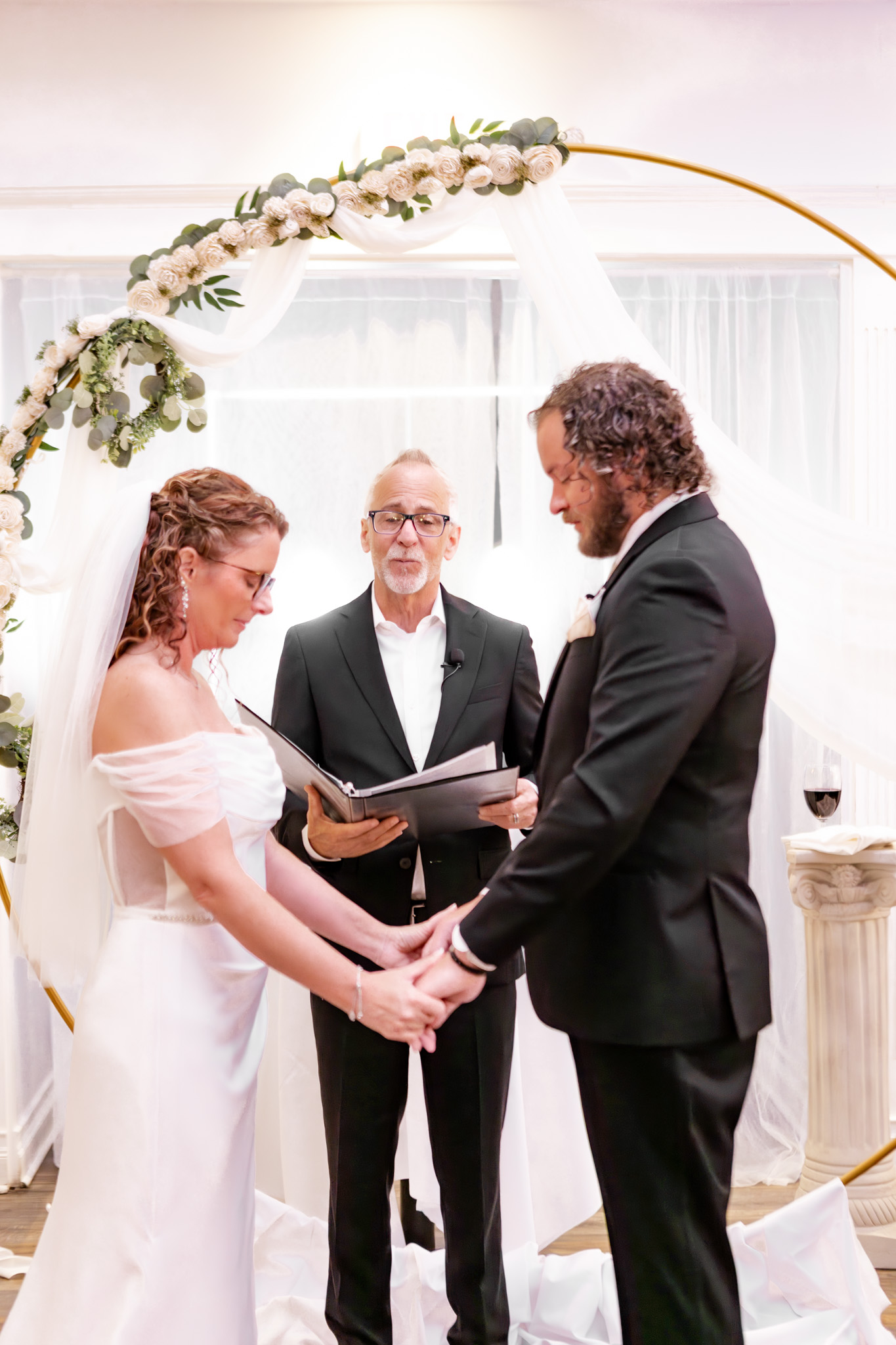 A bride and groom hold hands and face each other during their wedding ceremony, with an officiant behind them. They stand beneath a circular floral arch, all dressed formally—a moment as beautifully designed as an Elementor layout.